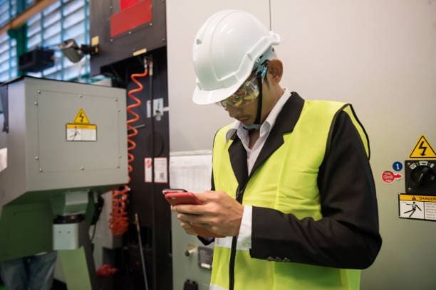 fire detection 4 Busy factory Asian engineer manager checking manufacturing project plan from 2 smartphone devices near production machine with safety warning symbol.
